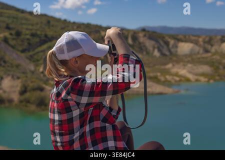 Frau in einem karierten Hemd und einer weißen Baseballmütze, die durch ihr Kameraobjektiv fokussiert und den ruhigen blauen See und die umliegenden trockenen Berge auf einem s dokumentiert Stockfoto