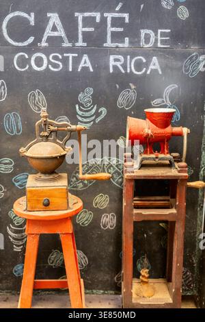 Vintage Kaffeemühle auf Holzständern vor der Tafel mit der Aufschrift „Café de costa rica“ mit Kaffeebohnenkritzeleien; rustikales Café-Dekor, warme Töne. Stockfoto