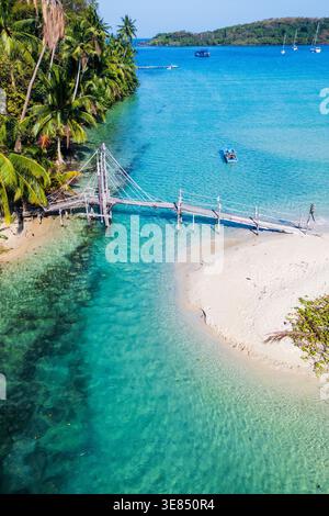 Blick aus der Vogelperspektive auf den tropischen Strand mit klarer türkisfarbener Lagune, Palmen, hölzerne Seilbrücke über flaches Wasser, kleine Boote und üppige Inselküste u Stockfoto