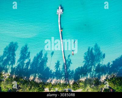 Blick von oben auf einen langen Pier, der sich ins türkisfarbene Meer erstreckt, mit kleinen Booten in der Nähe und Palmenschatten entlang einer tropischen Sandküste. Stockfoto