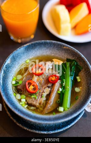 Schüssel asiatische Nudelsuppe mit geschmortem Rindfleisch, roten Chili-Scheiben, Zwiebeln und Pak Choy auf dunklem Tisch, mit Orangensaft und Obstteller im Hintergrund Stockfoto