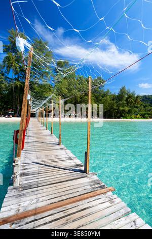Holzpier, der sich über türkisfarbenes Meer zum tropischen Inselstrand mit Palmen, klarem blauem Himmel und schimmernden Wolken erstreckt; Reiseurlaubskonzept, friedlich Stockfoto