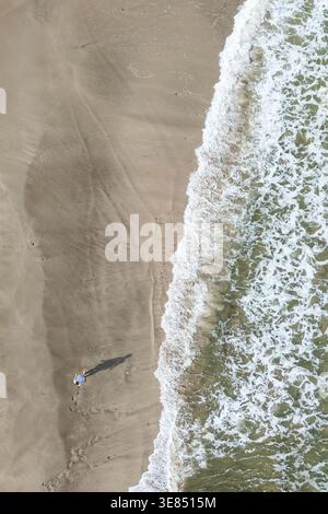 Aus der Vogelperspektive auf den Sandstrand mit einer einsamen Person, die langen Schatten wirft, strukturierte Surflinien und Küstenlinien, minimale Sommerreise im Hintergrund Stockfoto