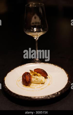 Feines Dining Dessertteller mit Sahnequenelle, karamelisierten Fruchtscheiben und Sauce Nieseln, mit leerem Weinglas im Hintergrund, stimmungsvollem, schwachem Licht, Sch Stockfoto