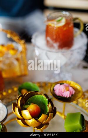 Luxuriöser thailändischer Nachmittagstee mit goldenen Lotus-Snackbechern, bunten Desserts und einem Eisgetränk in einem Glasbecher auf kunstvollen Tabletts, weichem Background Stockfoto