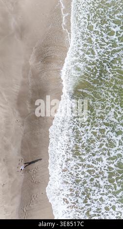 Aus der Vogelperspektive auf den Sandstrand mit einer einsamen Person, die langen Schatten wirft, strukturierte Surflinien und Küstenlinien, minimale Sommerreise im Hintergrund Stockfoto