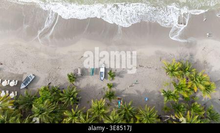 Blick aus der Vogelperspektive auf den tropischen Strand mit Meereswellen, Palmen und kleinen Booten auf Sand, minimale Menschen, sonnige Küstenurlaubszene. Stockfoto