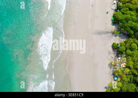 Blick von oben auf den tropischen Strand mit türkisfarbenen Wellen, breiter Sandstrand und üppig grünem Küstenwald; ein paar kleine Figuren und Sonnenschirme VI Stockfoto