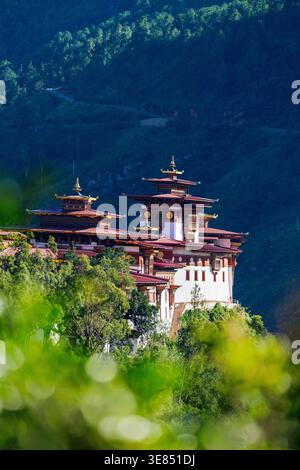 Traditionelles bhutanisches Dzong-Kloster mit gestuften roten Dächern auf einem bewaldeten Hügel vor nebeligen Bergen, heiterer buddhistischer Architektur und Natur-la Stockfoto