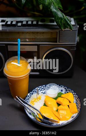 Mango-Reisdessert mit Kokosnusscreme und Gabeln neben Mango-Smoothie, vor einer Vintage-Boombox gesetzt; stimmungsvolles tropisches Stillleben. Stockfoto
