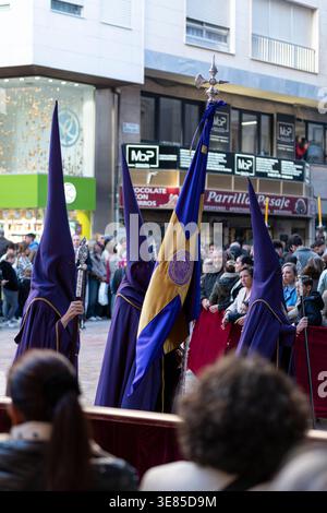 Almeria, Spanien - 30. März 2026: Pönitenten in violetten Gewändern und spitzen Kapuzen tragen während einer Karwochenprozession ein Prozessionsbanner als Zuschauer g Stockfoto