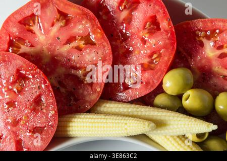 Eine hochschwingende Nahaufnahme saftiger Tomatenscheiben mit Salz und Pfeffer, serviert mit eingelegtem Babymais und grünen Oliven. Helles, natürliches Licht h Stockfoto