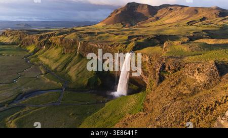 Aus der Vogelperspektive des Wasserfalls Seljalandsfoss, der bei Mitternachtssonne von grünen Klippen in die isländische Landschaft eintaucht, eingerahmt von zerklüfteten Bergen Stockfoto