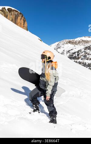 Snowboarderin in Helm und Schutzbrille, die auf einem hellen, verschneiten Hang in den Pyrenäen steht, ihr Brett hält und den klaren blauen Himmel genießt Stockfoto