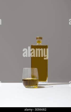Minimales Stillleben mit einer quadratischen Flasche goldenem Olivenöl und einem kleinen Verkostungsglas auf einer weißen Oberfläche, weichem Licht und neutralem Hintergrund, Elegane Stockfoto