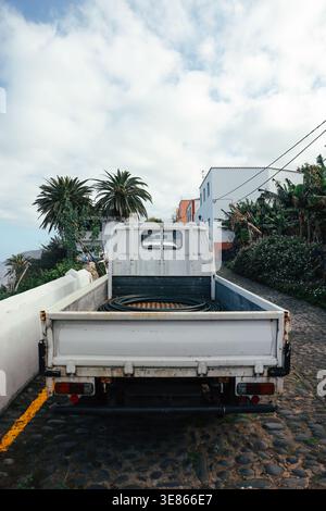 Rückansicht eines weißen Pickup-Trucks, der auf einer Kopfsteinpflasterstraße in einem ruhigen Dorf La Palma, den Kanarischen Inseln, mit Palmen, Häusern und einer bewölkten Ske geparkt ist Stockfoto