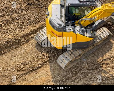 Draufsicht eines gelben Baggers auf Raupenfahrwerk, der auf einer Erde- und Kiesbaustelle arbeitet, mit Kabine, Hydraulikarm und strukturiertem Boden Stockfoto