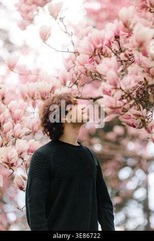 Nachdenklicher junger Mann, der unter blühenden rosafarbenen Magnolienzweigen steht und in einem hellen Frühlingsgarten auf weiche Blätter blickt und eine ruhige, romantische Atmosphäre schafft Stockfoto