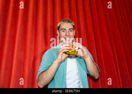 Fröhlicher junger Mann mit kurzen blonden Haaren hält einen frischen Burger und lächelt in einem Studio vor einem leuchtend roten Vorhang Stockfoto