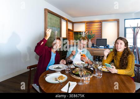 Freunde, die ein lebhaftes mediterranes Essen an einem gemütlichen Restauranttisch genießen, Paella und Pasta essen, Wein trinken, zusammen lachen und Telefone checken Stockfoto