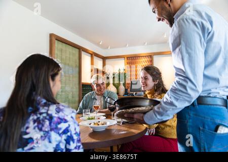 Gruppe von Freunden, die mediterrane Speisen in einem gemütlichen Restaurant genießen, während ein Kellner ein heißes Gericht serviert. Warme Lifestyle-Szene mit Lachen, Weinglas Stockfoto