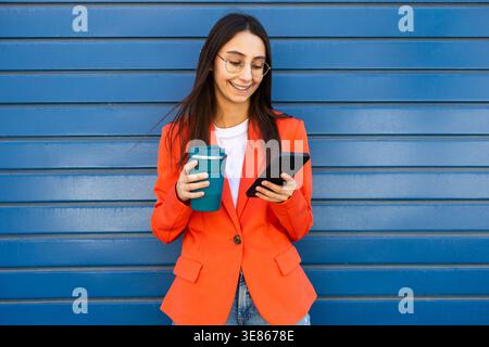 Eine lächelnde Frau in einer stylischen orangefarbenen Jacke hält eine wiederverwendbare Kaffeetasse, während sie ihr Smartphone überprüft, und steht vor einer blauen Wand. Sie verkörpert Moder Stockfoto