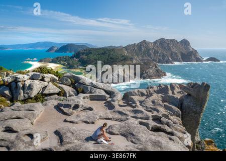 Frau sitzt auf sonnendurchfluteten Granitfelsen mit Blick auf das türkisfarbene Meer und die zerklüftete Küste der Cies-Inseln, Spanien, mit Stränden, Wellen und Inseln auf der Stockfoto