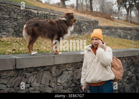 Junge Frau in warmer Jacke und Mütze geht draußen, während sie mit einem Smartphone spricht, während ein australischer Schäferhund auf einer Steinmauer in einem Park steht. Stockfoto