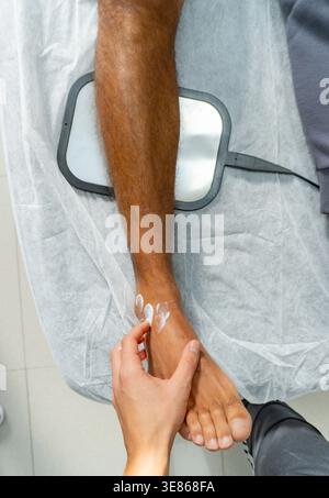 Oben Nahaufnahme einer Hand eines Therapeuten, die vor der Behandlung mit Rehabilitationsgeräten Creme auf den Knöchel des Patienten aufträgt, wobei Hautpflege und Schmerzreaktionen hervorgehoben werden Stockfoto