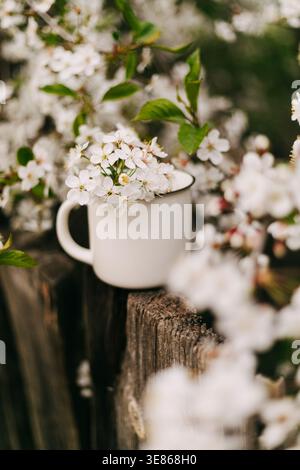 Traumhaftes Stillleben im Frühling mit zarten weißen Blüten in einem einfachen Emaillebecher auf einem rustikalen Holzzaun, umgeben von weichem Bokeh und Natur Stockfoto