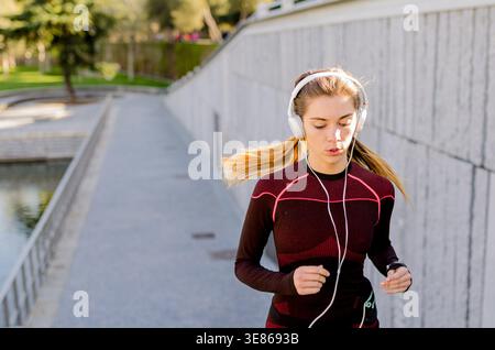 Junge Frau mit Kopfhörern und Sportbekleidung joggt auf einem sonnigen Weg in einem Stadtpark. Ihr fokussierter Ausdruck spiegelt Entschlossenheit und Hingabe wider Stockfoto