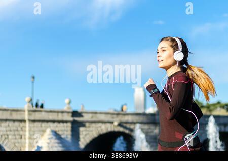 Frau in Sportswear joggt in einem Stadtpark, hört Musik über Kopfhörer und trägt eine Smartwatch. Der Hintergrund zeigt eine steinbrücke Stockfoto