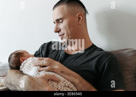 Ein kaukasischer Mann hält ein neugeborenes Mädchen in einer Decke. Das Baby hat dunkle Haare und schaut auf den Mann. Sie sind drinnen, mit einem gemütlichen atmos Stockfoto