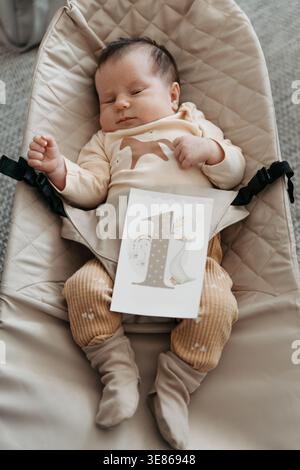 Ein schlafendes Mädchen in einem beigefarbenen Outfit liegt in einer Babywippe. Sie hält eine Karte mit der Nummer eins, die ihr Alter angibt. Weiche, neutrale Farben dominieren Stockfoto