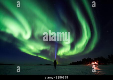 Leuchtend grüne aurora borealis schwingt über einem sternenklaren Winterhimmel über einem gefrorenen See in Finnland, während ein einsamer Wanderer nahe der Ferne eine Scheinwerferlampe strahlt Stockfoto