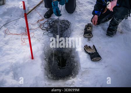 Eisfischen in Finnland mit einem Winternetz, das durch ein Loch in einem gefrorenen See gezogen wird. Nahaufnahme von Händen, Handschuhen und Ausrüstung auf Schnee, die traditionelles Outdo zeigt Stockfoto