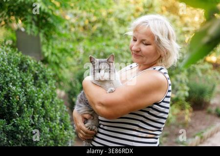 Glückliche Seniorin umarmt ihre graue Katze im sonnigen Garten. Stockfoto