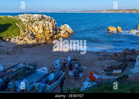 Mehrere kleine Fischerboote werden an einem Sandstrand gezogen, wo Fischer mit Netzen und Ausrüstung arbeiten, eingerahmt von einem großen Felsvorsprung und dem blauen Ozean Stockfoto