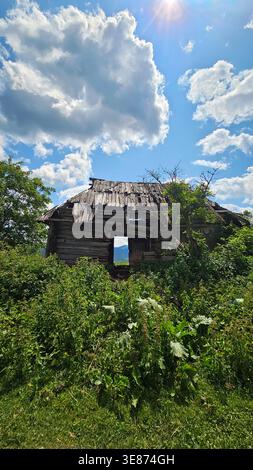 Verlassenes Holzhaus in Mountain Meadow. Altes, ruiniertes Holzhaus, das auf bewachsenem Gras unter einem hellblauen Himmel steht und große Wolken in einer Ferne hat Stockfoto