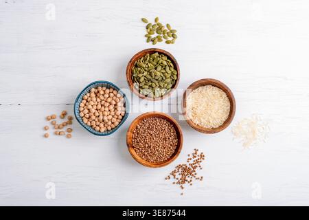 Getreide und Samen in Schüsseln auf weißem Holztisch. Gesundes Lebensmittelkonzept. Reis, Kichererbsen, Buchweizen und Kürbissamen. Draufsicht, flach. Stockfoto