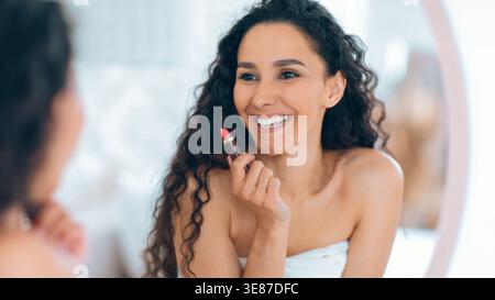 Frau trägt Lippenstift vor dem Spiegel auf, während sie im Morgenlicht zu Hause lächelt Stockfoto
