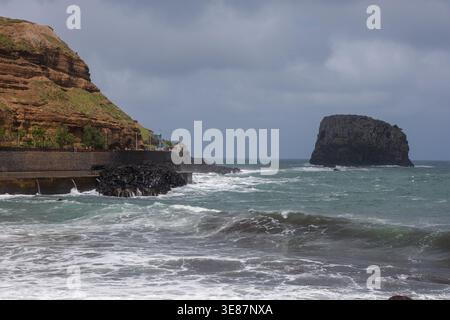 Bewölkter Himmel hängt über Wellen, die gegen dunkle vulkanische Felsen und eine Meeresmauer unter ockerfarbenen Terrassenklippen krachen, mit Ilheu da Ribeira da Janela ris Stockfoto