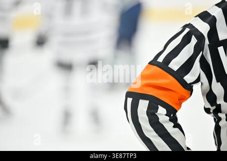 Eishockey-Schiedsrichter in gestreifter Uniform mit orangefarbenem Armband während eines Spiels, mit unscharfen Spielern im Hintergrund auf der Eisbahn. Stockfoto
