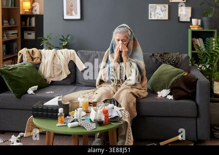 Ältere kaukasische Frau niest in Gewebe, während sie zu Hause in eine Decke gewickelt sitzt und saisonale Grippe oder Erkältung zeigt. Wide Shot, passende Anzeigen für das Gesundheitswesen, Apothekenkampagnen, krankheitsbedingte Inhalte Stockfoto