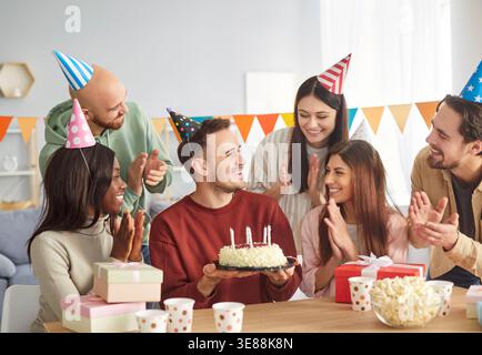 Fröhliche Gruppe von verschiedenen Freunden mit Partyhüten, die zu Hause mit Kuchen und Geschenken ihren Geburtstag feiern. Stockfoto