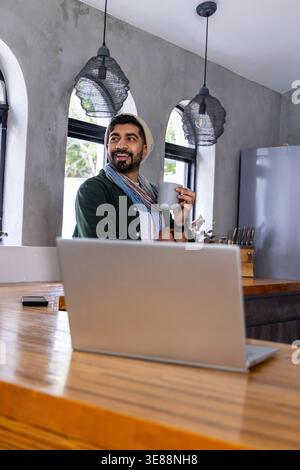 Indischer Mann, der hinter einer Holztheke in der modernen Küche steht und einen grauen Becher hält und einen Laptop benutzt Stockfoto
