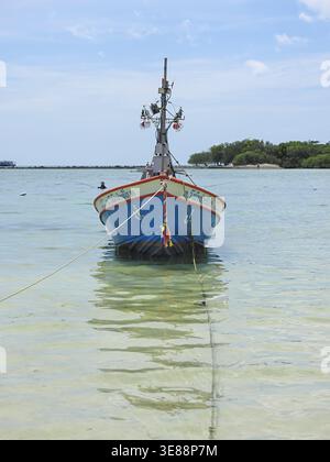 Blaues Holzboot, das im flachen, kristallklaren tropischen Meer mit einer fernen Insel und hellem Himmel vertäut, erinnert an friedliche Reisen und traditionelles Fischleben Stockfoto