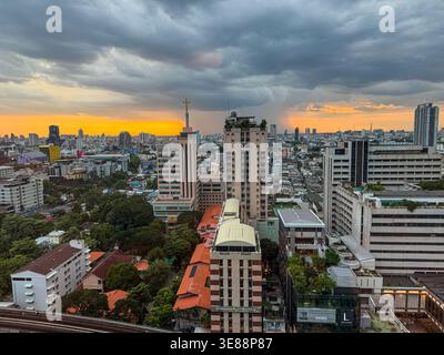 Dynamisches Stadtbild von Bangkok in der Dämmerung, das städtische Gebäude, Grünflächen, eine erhöhte Bahnlinie und einen entfernten Regenschauer im Kontrast zu einem Gold zeigt Stockfoto