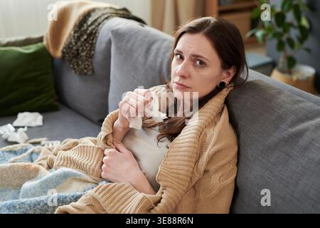 Porträt einer kaukasischen jungen erwachsenen Frau, die auf dem Sofa ruht, Gewebe hält und in die Kamera schaut, zeigt Erkältung oder Grippe Erholung. Nützlich für Anzeigen im Gesundheitswesen, krankheitsbedingte Abwesenheit, häusliche Behandlungskampagnen Stockfoto