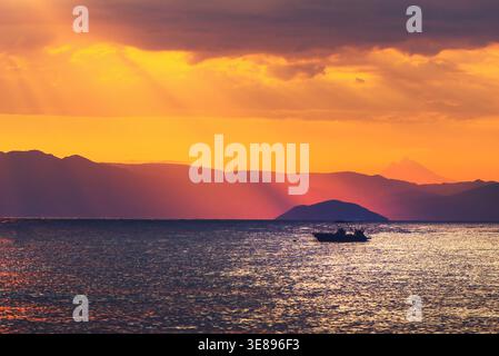 Silhouette eines Bootes auf ruhigem Meer bei lebendigem Sonnenuntergang, mit mehrschichtigen Bergketten im Hintergrund und dramatischen Sonnenstrahlen durch die Wolken. Wählen Sie Aus Stockfoto